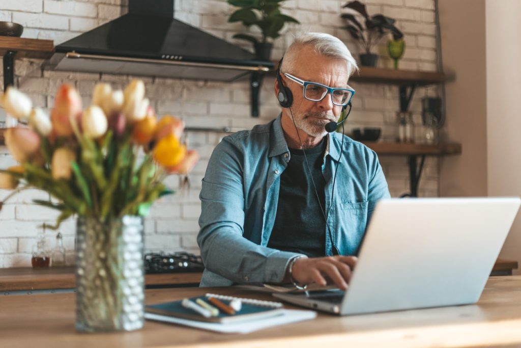 Older man using a laptop and wearing a headset in his kitchen